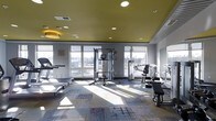Located at 3001 Village Market Pl in Morrisville, NC, this fitness center offers a well-appointed, single-floor space designed to support a comprehensive workout experience. The room features a bold yellow ceiling accented by recessed lighting, creating a bright and energizing atmosphere that complements the abundant natural light streaming in through large floor-to-ceiling windows along the perimeter walls. These expansive windows provide open views of the surrounding area, contributing to an airy, motivating environment. The space is equipped with a thoughtfully curated selection of exercise machines, including a cable crossover station, strength training equipment, and cardio machines such as a treadmill. The flooring consists of a patterned, multi-toned carpet that adds visual interest while providing comfort underfoot. Mirrored walls further enhance the sense of openness and allow users to monitor their form during workouts. The neutral gray walls, combined with the vivid ceiling and natural light, strike a balance between functional design and modern aesthetics, making this fitness center a well-rounded facility for residents and visitors in the Village Market Place community.