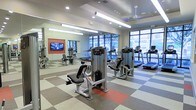 This fitness center offers a comprehensive workout environment designed to accommodate a wide range of exercise needs. The space is outfitted with a selection of modern strength-training machines, including a prominent multi-function weight machine at the center of the floor, alongside a variety of cardio equipment such as treadmills positioned near large floor-to-ceiling windows. The expansive windows flood the interior with abundant natural light and provide open views to the outdoors, creating an inviting and energizing atmosphere. Full-length mirrors line one wall, visually expanding the space and allowing users to monitor their form during workouts. A flat-screen television is mounted within the mirrored section, adding an entertainment element to the exercise experience. The flooring features a distinctive checkered pattern using multi-toned rubber tiles in grey, beige, and red, offering both durability and visual interest. Overhead, a combination of recessed lighting and linear fluorescent fixtures ensures thorough, even illumination throughout the room. A ceiling fan contributes to air circulation and comfort during intense workouts. The overall design is clean, functional, and modern, making this fitness center a well-appointed facility suited for daily use.