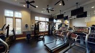 This fitness center is a thoughtfully designed, single-floor facility that offers a comprehensive workout environment for users of all fitness levels. The space features classic architectural detailing, including crown molding, a two-tone wall treatment with a rich burgundy wainscoting below and neutral warm tones above, and large multi-pane windows fitted with wooden blinds that allow generous natural light to fill the room.

The cardio section is well-appointed, housing multiple treadmills, elliptical trainers, upright and recumbent stationary bikes, and a stair climber, providing a wide variety of aerobic exercise options. A dedicated strength training area includes a multi-station cable machine and a full dumbbell rack, catering to resistance and weight training needs. A rowing machine rounds out the equipment offerings.

Multiple ceiling fans distributed throughout the space ensure comfortable air circulation during workouts. Several wall-mounted flat-screen televisions are positioned for easy viewing from most equipment stations, enhancing the exercise experience. Full-length mirrors along one wall visually expand the space and allow users to monitor their form. Rubber flooring covers the entire workout area, providing safety and durability. A defibrillator is visibly mounted on the wall, reflecting a commitment to user safety. A glass-panel door connects the fitness center to an adjacent room, adding to the facility's overall accessibility.