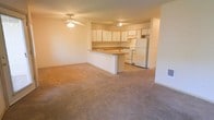 This single-floor space showcases an open-concept layout that seamlessly connects the living area and kitchen, creating a sense of openness and ease of flow. The generously sized living area is finished with wall-to-wall carpeting in a neutral tone, providing a warm and comfortable underfoot feel. A ceiling fan with a light fixture is centrally positioned overhead, offering both air circulation and ambient lighting to the space.

The kitchen is thoughtfully designed with an abundance of white upper cabinetry that extends to the ceiling, maximizing storage capacity. White appliances, including a refrigerator and a range, are neatly integrated into the layout. A peninsula-style breakfast bar separates the kitchen from the living area, offering a practical surface for casual dining or food preparation while maintaining the open feel of the space.

The kitchen flooring transitions to a hard surface, clearly delineating it from the carpeted living zone. The walls throughout are finished in a soft, neutral tone that reflects light and contributes to the bright, airy ambiance of the space. The overall design is clean, functional, and well-suited for everyday living.