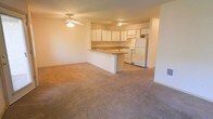 This single-floor space showcases an open-concept layout that seamlessly connects the living area and kitchen, creating a sense of openness and ease of flow. The generously sized living area is finished with wall-to-wall carpeting in a neutral tone, providing a warm and comfortable underfoot feel. A ceiling fan with a light fixture is centrally positioned overhead, offering both air circulation and ambient lighting to the space.

The kitchen is thoughtfully designed with an abundance of white upper cabinetry that extends to the ceiling, maximizing storage capacity. White appliances, including a refrigerator and a range, are neatly integrated into the layout. A peninsula-style breakfast bar separates the kitchen from the living area, offering a practical surface for casual dining or food preparation while maintaining the open feel of the space.

The kitchen flooring transitions to a hard surface, clearly delineating it from the carpeted living zone. The walls throughout are finished in a soft, neutral tone that reflects light and contributes to the bright, airy ambiance of the space. The overall design is clean, functional, and well-suited for everyday living.