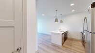 This 1-bedroom, 1-bathroom unit in McCordsville, Indiana offers a thoughtfully planned layout across a single floor. The open-concept kitchen and living area serves as the heart of the home, featuring light wood-style flooring, crisp white walls, and recessed lighting throughout. The kitchen is well-appointed with dark espresso cabinetry, white quartz countertops, a peninsula with an undermount sink, stainless steel appliances—including a refrigerator, electric range, and microwave—and a light blue subway tile backsplash. Stylish pendant lights hang above the peninsula, adding a modern touch. The living area provides a bright, open canvas with ample natural light from large windows. The bedroom is carpeted for comfort, includes a ceiling fan, and offers access to a generously sized walk-in closet outfitted with wire shelving systems. The full bathroom features dark wood vanity cabinetry, white countertops, and a fully tiled walk-in shower with a handheld showerhead. A dedicated laundry area houses a stacked washer and dryer unit alongside a water heater, keeping utilities efficiently organized. Clean lines and neutral tones throughout create a cohesive, contemporary atmosphere.