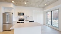 Located at 30 Rue Jos-Montferrand in Gatineau, QC, this single-floor unit offers a thoughtfully designed layout that maximizes its compact footprint. The space features a striking exposed concrete ceiling throughout the main living and kitchen areas, lending an industrial-modern character that contrasts elegantly with the bright white walls and light hardwood-style flooring found throughout.

The open-concept kitchen serves as a central focal point, equipped with stainless steel appliances including a refrigerator, electric range, and built-in microwave. Crisp white cabinetry extends to the ceiling, providing ample storage, while a large central island with a white quartz-style countertop offers additional workspace and informal seating. A stainless steel dishwasher and sink complete the functional layout.

The bedroom area benefits from a large floor-to-ceiling window that frames panoramic city views of Gatineau, filling the space with abundant natural light. A sliding barn-style door and built-in storage solutions are integrated efficiently into the hallway.

The bathroom is finished with neutral beige tile, featuring a full soaking tub with shower, a white vanity, and recessed lighting. Clean lines and a restrained, neutral palette unify the entire unit, creating a cohesive and contemporary living environment.