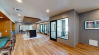 This thoughtfully designed single-floor workspace blends modern aesthetics with highly functional amenities. Warm hardwood flooring runs throughout the open main corridor, connecting the various zones of the space seamlessly. A row of glass-enclosed private work pods lines one wall, each equipped with a desk, ergonomic seating, and views to the outdoors — ideal for focused, individual work. The open lounge area features a striking built-in banquette accented by a bold geometric mural in teal, cream, and charcoal tones, complemented by sleek side tables. Adjacent to the lounge is a well-appointed kitchenette with vibrant teal cabinetry, geometric tile backsplash, dark stone countertops, and a large island topped with a light stone surface and green upholstered bar stools beneath a warm wood ceiling panel with pendant lighting. Two dedicated conference rooms offer textured wall coverings, curated artwork, and seating for small groups — one fitted with globe pendant lights and a rectangular table, the other with a round table and blue task chairs. Throughout the space, colorful accent furniture, framed artwork, and ample natural light from large windows create a vibrant, professional, and welcoming atmosphere.