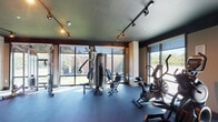 Located at 1700 Marinas Edge Way in Louisville, Kentucky, this fitness center is a thoughtfully designed, single-floor facility built to support a full range of workout routines. The space features a dark, industrial-style ceiling with exposed ductwork and track lighting throughout, creating a modern, energetic atmosphere well-suited for exercise.

Large floor-to-ceiling windows line multiple walls of the facility, flooding the interior with abundant natural light and offering views of the surrounding greenery and outdoor areas. The flooring consists of durable rubber gym matting, providing both safety and comfort underfoot.

The cardio zone is well-appointed with treadmills, stationary bikes, and an elliptical trainer, all positioned to take advantage of the natural light and outdoor views. The strength training area features a multi-station cable machine, a dedicated free weights section with a tiered dumbbell rack, and a variety of resistance and functional training tools including medicine balls, resistance rings, and foam rollers organized neatly on a storage rack. A wall-mounted flat-screen television is available in the free weights area.

An additional room houses treadmills and a Peloton-style stationary bike, accessible through a glass-panel interior door. The overall ambiance is sleek and functional, making this fitness center a comprehensive resource for residents seeking convenient, on-site exercise facilities.