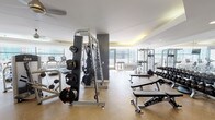 Located at 301 E North Water St in Chicago's Streeterville neighborhood, this fitness center offers a thoroughly equipped workout facility designed to accommodate a full range of exercise needs. The space is laid out across a single level, with distinct zones dedicated to cardiovascular training and strength work.

The cardio area features a row of modern treadmills, elliptical trainers, and stair climbers aligned along floor-to-ceiling windows, providing unobstructed views of the Chicago skyline and waterfront. A mounted flat-screen television accompanies the cardio zone, enhancing the workout experience. The natural light from the expansive windows creates a bright and energizing atmosphere throughout the facility.

The strength training section is well-stocked with cable machines, multi-functional weight stations, adjustable benches, and a comprehensive rack of Matrix-branded dumbbells. The layout allows ample space for movement between equipment. Large mirrors along one wall visually expand the space and assist with form during exercises.

Near the entrance, built-in cabinetry with open shelving stocks fresh towels, and a water station is conveniently positioned for use. The flooring throughout is a durable speckled rubber composite, and a tray ceiling with warm accent lighting adds a polished architectural detail to the overall design.