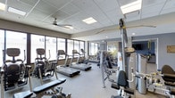 This fitness center is designed to support a comprehensive workout experience within a single-level layout. The space is outfitted with a solid selection of cardio equipment, including multiple treadmills and an elliptical trainer, positioned along a wall of large horizontal windows that allow natural light to fill the room and provide views to the exterior. A ceiling fan overhead aids in air circulation, contributing to a comfortable environment during workouts.

On the strength training side, the facility includes cable weight machines, a dedicated bench press station, and a dumbbell rack, offering users a range of resistance training options. Mirrored walls on one side of the room visually expand the space and allow users to monitor their form during exercises. Decorative vertical accents on the mirrored wall add a subtle design touch to the otherwise utilitarian aesthetic.

The flooring consists of durable, light-toned gym-appropriate material suited for high-traffic use. Recessed and panel lighting in the drop ceiling supplements natural light, ensuring the space remains well-lit throughout the day. The overall ambiance is clean, modern, and functional.