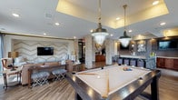 This single-floor amenity space is a well-appointed game room and social lounge designed to encourage community and leisure. At its center sits a full-size pool table, illuminated by striking brass and industrial-style pendant lights suspended from a coffered ceiling, setting an inviting tone throughout the room. Wood-look tile flooring runs seamlessly across the space, tying together the various activity zones with a cohesive, contemporary aesthetic.

The lounge area features plush leather sofas, an accent armchair, and a shag area rug, all positioned around a flat-screen TV mounted against a textured chevron-patterned accent wall. Large floor-to-ceiling windows flood this area with natural light and offer views of the surrounding outdoors, while sheer drapes add a soft, layered finish.

A granite-topped bar counter with upholstered round stools serves as a social hub, flanked by a wall of wood-toned tile and framed by globe pendant lighting. Behind it, multiple flat-screen TVs and decorative mirrors create an entertainment-focused backdrop. A dedicated beverage and refreshment station with rich wood cabinetry, a draft tap, and refrigeration units is tucked nearby, accented by stone-clad structural columns. A cozy bistro table nook and a cue rack mounted between tile columns round out this thoughtfully designed communal space.