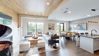This thoughtfully designed single-floor space seamlessly integrates a living room, dining room, and kitchen into one cohesive open-plan layout. The living area is anchored by a stunning wood-planked tray ceiling that adds warmth and architectural character, complemented by a sleek modern fireplace set into a matching wood accent wall. A grand piano occupies a prominent corner beneath the ceiling feature, adding a refined focal point. Large windows and glass balcony doors flood the space with natural light while offering lush outdoor views. Rich hardwood flooring flows consistently throughout, tying the zones together. The kitchen is outfitted with floor-to-ceiling light grey shaker cabinetry, dark veined stone countertops, a bold matching backsplash, and stainless steel appliances. A generous waterfall island with an undermount sink and black pendant lighting provides both functionality and a striking visual centerpiece. The dining area accommodates a large dark wood table with woven accent chairs, bathed in natural light from full-height windows. Neutral tones, warm wood textures, and contemporary furnishings throughout create a sophisticated yet inviting ambiance that balances comfort with modern elegance.