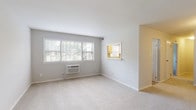 This single-floor residence offers a clean, neutral aesthetic throughout, with light gray walls, white trim, and plush wall-to-wall carpeting that lends a comfortable, cohesive feel to every room. The layout, as seen in the dollhouse view, flows efficiently from a spacious living area through a central corridor connecting the kitchen, bathroom, and additional rooms.

The living area is generously sized and filled with natural light from a large multi-pane window fitted with blinds, which offers views of surrounding greenery. A wall-mounted air conditioning unit ensures year-round climate comfort. A pass-through window connects the living space to the kitchen, adding an open, communicative feel between the two areas.

The kitchen is well-equipped with white shaker-style cabinetry, stone-look laminate countertops offering ample workspace, a stainless steel sink beneath a window, and space for appliances including a gas range. The bright, functional layout makes the kitchen both practical and inviting.

Additional carpeted rooms provide versatile space, each featuring windows that bring in natural light and wall AC units. The bathroom is finished in a neutral palette with white fixtures, including a toilet and sink, complemented by soft gray walls.

Overall, the space presents a bright, clean, and functional environment with consistent finishes throughout.