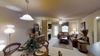 This single-floor residence offers an inviting, open-concept layout that seamlessly connects the dining and living areas, creating a cohesive and functional living environment. The dining area is anchored by a classic pendant light overhead and features a traditional wooden dining set, complemented by lush greenery that adds a natural, homey touch.

The living room is generously proportioned, centered around a built-in fireplace that serves as a focal point, with a wall-mounted television positioned above. Rich, earth-toned leather sofas and a patterned area rug over neutral carpeting contribute to the warm, comfortable ambiance throughout. A ceiling fan with a light fixture enhances airflow and illumination.

Natural light fills the space through a glass-paneled door and a window dressed with decorative valances, offering a view to the exterior. A decorative sideboard with accent pieces, framed artwork, and coordinated accessories add character and depth to the walls. The overall aesthetic is traditional and welcoming, with warm neutral tones, layered lighting, and carefully arranged furnishings creating a cohesive, lived-in atmosphere.