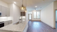 This thoughtfully designed two-bedroom, two-bathroom residence combines modern aesthetics with functional living across a single floor. The open-concept kitchen serves as the heart of the home, featuring granite countertops, a large center island with an undermount sink, gray shaker-style cabinetry, stainless steel appliances, a tile backsplash, and striking geometric pendant lighting. Wood-look flooring flows seamlessly from the kitchen into the living area, which is bathed in natural light from large windows and accented by a ceiling fan overhead. Built-in shelving adds a practical decorative touch near the main living space. The primary bedroom is generously sized and connects to a spacious en-suite bathroom with a dual-sink granite vanity, marble-look tile flooring, and a walk-in shower. A large walk-in closet with multi-level shelving and hanging rods provides exceptional storage. The second bathroom features the same elegant marble-look tile, a soaking tub with a tiled surround, and a granite vanity. The second bedroom offers wood-look flooring and a bright window with outdoor views. Clean white walls and recessed lighting throughout create a bright, cohesive ambiance across the entire residence.