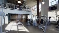 This two-story fitness facility showcases an industrial-inspired design with soaring double-height ceilings that create an expansive, open atmosphere. Exposed ductwork runs overhead, complementing the raw architectural aesthetic, while large windows along the upper walls flood the space with abundant natural light, casting dynamic shadows across the rubber-floored workout area below.

The main floor is equipped with a range of professional-grade strength training machines, including cable and resistance units arranged to allow comfortable circulation between stations. The dark rubber flooring provides both durability and safety underfoot. Glass-paneled partitions along one side of the main floor divide the space into functional zones while maintaining visual openness throughout.

A mezzanine level accessible via a staircase with metal railings overlooks the main workout floor, offering additional exercise space—visible from below are cardio machines positioned to take advantage of the elevated views. Industrial pendant lighting fixtures illuminate both levels consistently. The overall ambiance is modern and functional, combining the character of an industrial loft with the practical demands of a well-equipped fitness center.