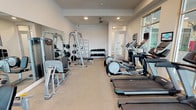 Located at 5406 Middle Fiskville Rd in Austin, Texas, this fitness center offers a comprehensive workout environment designed to accommodate a wide range of exercise needs. The space is housed on a single floor and is characterized by neutral-toned walls, recessed lighting, and a ceiling fan that promotes comfortable air circulation throughout the room.

Large, framed windows line one side of the facility, allowing generous natural light to fill the space and providing views to the exterior. The open layout is organized to maximize usability, with cardio equipment — including multiple treadmills, elliptical trainers, and a stationary bike — arranged along one side of the room, each unit equipped with individual monitors for entertainment during workouts.

The strength training area features a multi-station cable and weight machine, a dedicated dumbbell rack stocked with a full range of free weights, and adjustable benches for varied exercises. Floor-to-ceiling mirrors along the walls enhance the sense of space and allow users to monitor their form. A shelving unit stocked with fresh towels adds a convenient, amenity-focused touch.

A glass-paneled door leads to an adjoining room, and a water station is available within the space. The carpeted flooring, clean finishes, and organized equipment arrangement contribute to a professional and welcoming atmosphere suited for daily fitness routines.