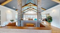 Located at 170 Cycle Dr in Boiling Springs, SC, this single-floor clubhouse is defined by its striking architectural character and open, airy layout. The centerpiece of the space is a dramatic vaulted ceiling accented with exposed wood beams painted in a deep navy-blue finish, lending a modern yet rustic charm throughout. Prominent stacked-stone columns rise from floor to ceiling, framing the open-concept interior and serving as bold structural focal points.

Warm hardwood flooring flows consistently across the main gathering areas, complementing the neutral palette and coastal-inspired décor. The lounge area is generously proportioned, featuring large sliding glass doors topped with expansive transom windows that flood the space with natural light and offer unobstructed views of the outdoor pool and patio area beyond.

A dedicated dining area provides a casual seating space anchored by a wood table and mixed-material chairs, positioned near the stone columns. The fully equipped kitchen features dark cabinetry, granite countertops, stainless steel appliances, a large central island with seating, and decorative pendant lighting, making it well-suited for gatherings and events.

Additional amenities, including what appears to be a fitness area, are accessible from within the main space. Overall, the clubhouse presents a welcoming, upscale atmosphere with cohesive design elements throughout.