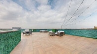 Located atop 20 Grand Ave in Englewood, NJ, this rooftop terrace is a well-appointed outdoor amenity space designed for relaxation and social gatherings. The terrace is finished with wood-look porcelain tile pavers across a generous open-air footprint, complemented by lush artificial turf sections that add a vibrant, park-like element to the environment.

The space is thoughtfully divided into distinct activity zones. A dedicated lounge area features wicker seating arrangements with plush white cushions, ideal for casual conversation or unwinding. A separate dining and grilling station is equipped with an outdoor grill and table seating, making it well-suited for al fresco entertaining. The turf zone offers a more relaxed setting, furnished with a hammock, a circular rattan daybed, and bean bag-style seating — all oriented to take in the surrounding skyline and treetop views of Englewood and beyond.

Lush green hedge panels line the perimeter walls, providing both privacy and an aesthetically cohesive backdrop throughout the space. Overhead string lights crisscross portions of the terrace, creating a warm ambiance for evening use. Open sightlines to the sky and horizon give the terrace a spacious, airy character, while the varied seating arrangements ensure it can accommodate both intimate gatherings and larger social events.