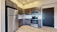 Located at 704 Eighth St in Niagara Falls, NY, this single-floor unit offers a thoughtfully designed interior with modern finishes throughout. The kitchen is a standout feature, showcasing rich dark gray shaker-style cabinetry — both upper and lower — paired with crisp light countertops accented by a marble-look tile backsplash. Stainless steel appliances, including a full-size refrigerator, electric range, and over-the-range microwave, complete the functional and stylish kitchen setup. Light wood-look flooring runs seamlessly throughout the unit, contributing to a cohesive and contemporary aesthetic. The open layout connects the kitchen to the adjacent living area, which benefits from natural light streaming in through a large multi-pane window with a black frame, offering views of the surrounding neighborhood. The bathroom is finished in clean, all-white tones and features a walk-in shower unit with built-in corner shelving, complemented by the same light flooring found throughout the space. Recessed lighting in the kitchen adds a polished, well-lit ambiance, while neutral wall tones provide a versatile backdrop throughout. The overall design reflects a clean, move-in-ready interior with modern upgrades suited for comfortable everyday living.