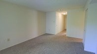 This single-floor space presents a clean, functional layout with a simple and practical design. The main room is generously sized, featuring wall-to-wall carpet in a neutral gray-beige tone that extends throughout the space, providing a consistent and cohesive look underfoot. The walls are finished in a light cream tone, contributing to a bright and open atmosphere, while the textured popcorn ceiling adds a classic touch overhead.

A flush-mount ceiling light fixture is visible in the hallway, offering modest illumination along the connecting corridor. The hallway leads to additional rooms, with at least two doors visible — suggesting access to separate spaces such as a bedroom or storage area, as well as an entry point. The layout appears straightforward, with a partial wall creating a natural division between the main room and the hallway, helping to define distinct zones within the space.

Ventilation is provided via a ceiling vent visible in the main area. The overall ambiance is understated and neutral, offering a blank canvas well-suited for a variety of furnishing and décor styles.