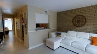This single-floor space presents a thoughtfully designed living area that blends contemporary and rustic elements. The room's most striking feature is a full-height exposed brick accent wall in warm earthy tones, providing texture and character to the space. Complementing this is a built-in fireplace framed by a clean white surround with a granite or stone hearth, serving as a functional and visual focal point.

The living area is furnished with a modern white sectional sofa and matching ottoman, adding a sleek, minimalist contrast to the more rustic brick backdrop. Colorful accent pillows introduce subtle pops of blue and yellow, breaking the neutral palette without overwhelming it.

The walls are finished in a soft warm cream tone, contributing to the bright and open feel of the space. Light-toned flooring further enhances the airy atmosphere. A hallway visible to one side suggests additional rooms beyond, with built-in shelving offering practical storage. The overall ambiance is clean, comfortable, and balanced, blending warm textures with modern furnishings for a welcoming interior environment.