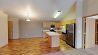 This single-floor space showcases an open-concept layout that seamlessly integrates the kitchen and living areas. The kitchen is well-appointed with light wood cabinetry that extends to the upper walls, providing generous storage throughout. Stainless steel appliances, including a refrigerator, electric range, and over-the-range microwave, lend a modern, functional character to the space. A central kitchen island with a light-toned countertop offers additional workspace and serves as a natural dividing point between the kitchen and the adjoining living area. A sink is positioned along the back counter, accompanied by additional cabinetry for ample storage. Warm hardwood flooring runs throughout the main area, transitioning to carpet at the living space edge, adding texture and comfort underfoot. The vaulted ceiling enhances the sense of openness, while a pendant light above the island and a ceiling-mounted fixture over the kitchen provide layered lighting. Neutral-toned walls complement the warm wood tones, creating a cohesive and inviting atmosphere throughout the space.