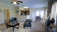 This single-floor residence offers an open-concept floor plan that seamlessly connects the living, dining, and kitchen areas into one cohesive and functional living space. The living room is anchored by plush wall-to-wall carpeting and features a ceiling fan overhead, crown molding details, and stylish furnishings including leather seating and a patterned area rug that adds a warm, inviting character to the space. The kitchen is positioned on a raised peninsula with dark wood cabinetry, stainless steel appliances — including a microwave and refrigerator — and a breakfast bar that provides a natural transition between the kitchen and the main living area. Adjacent to the living room, the dining area accommodates a full dining table and chair set, bathed in natural light streaming through a large sliding glass door that opens to the outdoors. The neutral, light-toned walls throughout create a bright and airy atmosphere, while the consistent flooring and open layout make the space feel expansive and easy to navigate. Crown molding and recessed lighting add refined architectural touches throughout, contributing to the overall polished and comfortable ambiance of the home.