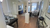 This single-floor residence offers an open and thoughtfully designed layout that seamlessly connects the living, dining, and entry areas into a cohesive living environment. Rich hardwood flooring flows throughout the space, lending warmth and a sense of continuity from room to room. Classic architectural details are evident throughout, including elegant crown molding and a distinctive white structural column that serves as a subtle divider between the dining and living areas without disrupting the open feel of the floor plan. The dining area features a dark wood table and chair set, positioned conveniently near the entryway. Built-in shelving units provide both display and storage options along the walls, adding functional character to the space. The living area is bathed in natural light, courtesy of large windows dressed with striped curtains, offering views of the surrounding greenery outside. A comfortable seating arrangement anchors the living space, complemented by a stylish arc floor lamp and area rugs that define each zone. A dark-toned dresser or media console adds a bold contrast against the light-colored walls. The overall ambiance is bright, airy, and inviting, with a classic yet contemporary aesthetic that balances comfort and style throughout.