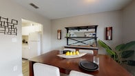 This single-floor residence offers a well-organized layout with a cohesive, neutral palette throughout. The living room serves as the heart of the home, featuring dark-toned hardwood-style flooring, a sliding glass door that floods the space with natural light, and direct access to a private outdoor patio. The open flow connects seamlessly to the dining area, which is highlighted by a rich wood dining table, upholstered chairs, and a decorative bar unit complete with wine storage and glass display shelving — a practical and stylish focal point.

The kitchen is efficiently designed with white cabinetry, light countertops, and full appliances including a coil-burner range with overhead ventilation and a full-size refrigerator. A pass-through view into the dining area reinforces the open feel of the shared living spaces.

The bedroom is a comfortable retreat, offering dark hardwood-style flooring, dual windows that invite natural light, and a sliding closet with built-in shelving. The full bathroom features a tiled tub-shower combination, a white vanity with countertop sink, and matching hardwood-style flooring, maintaining a clean and consistent aesthetic throughout the home.