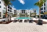Located at 3065 Champion Ring Rd in Fort Myers, Florida, this outdoor amenity courtyard is the centerpiece of a modern multi-story residential community. The space is defined by a large, resort-style swimming pool with a gracefully curved silhouette and crystal-clear turquoise water, serving as the focal point of the entire complex. The pool deck is finished in light-toned pavers and is generously appointed with sleek lounge chairs and shaded umbrella seating, providing ample space for relaxation under the Florida sun.

The courtyard is enclosed on multiple sides by a contemporary four-story building featuring a crisp white exterior, large windows, private balconies, and a clean, modern architectural aesthetic. Towering palm trees are strategically placed throughout the space, lending a lush, tropical ambiance that complements the sunny Southwest Florida setting. Tropical plantings and manicured garden beds add layers of greenery throughout.

A secondary courtyard area features a herringbone-patterned paver walkway flanked by low planters, tropical foliage, and a striking red sculptural accent piece, offering a calm, design-forward gathering space. Thatched market umbrellas, additional lounge seating, and thoughtful lighting complete the outdoor experience. The overall environment is open-air, bright, and designed to encourage an active, leisure-focused lifestyle.