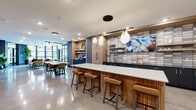 This single-floor community lounge at 215 E Prospect Ave in Mount Prospect, Illinois, showcases a thoughtfully designed, open-plan layout that seamlessly blends social and leisure spaces. Polished concrete flooring extends throughout, reflecting light and reinforcing the space's contemporary aesthetic.

The kitchen area serves as a central hub, anchored by a substantial island with a fluted wood base and crisp white countertop, surrounded by cognac leather barstools. Warm wood cabinetry, a textured tile backsplash, open shelving, and pendant globe lighting create an inviting, cafe-inspired atmosphere. A mounted television, built-in coffee station, and ample counter space make this area functional for gatherings.

Adjacent to the kitchen, a dining zone provides multiple seating configurations beneath sculptural pendant lighting. The lounge area features rich leather seating arranged around a coffee table, anchored by a patterned area rug. Floor-to-ceiling windows curve around this corner, flooding the space with natural light and providing direct access to an outdoor terrace.

A brick accent wall runs along one side, lending industrial character and housing a wall-mounted television, bar-height counter, and shuffle board table. A separate entry corridor features the same exposed brick, complemented by open metal shelving. The overall ambiance is modern, welcoming, and well-appointed.
