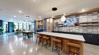 This single-floor community lounge at 215 E Prospect Ave in Mount Prospect, Illinois, showcases a thoughtfully designed, open-plan layout that seamlessly blends social and leisure spaces. Polished concrete flooring extends throughout, reflecting light and reinforcing the space's contemporary aesthetic.

The kitchen area serves as a central hub, anchored by a substantial island with a fluted wood base and crisp white countertop, surrounded by cognac leather barstools. Warm wood cabinetry, a textured tile backsplash, open shelving, and pendant globe lighting create an inviting, cafe-inspired atmosphere. A mounted television, built-in coffee station, and ample counter space make this area functional for gatherings.

Adjacent to the kitchen, a dining zone provides multiple seating configurations beneath sculptural pendant lighting. The lounge area features rich leather seating arranged around a coffee table, anchored by a patterned area rug. Floor-to-ceiling windows curve around this corner, flooding the space with natural light and providing direct access to an outdoor terrace.

A brick accent wall runs along one side, lending industrial character and housing a wall-mounted television, bar-height counter, and shuffle board table. A separate entry corridor features the same exposed brick, complemented by open metal shelving. The overall ambiance is modern, welcoming, and well-appointed.