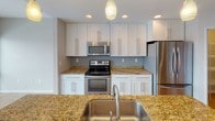This single-floor residence offers a thoughtfully designed layout with a clean, contemporary aesthetic throughout. The kitchen serves as a highlight of the space, showcasing white shaker-style cabinetry extending to the ceiling, warm gold-toned granite countertops, and a full suite of stainless steel appliances including a range, over-the-range microwave, and refrigerator. A central island with an undermount sink and pendant lighting above provides additional workspace and a natural gathering point. Recessed lighting and light wood-style flooring complete the kitchen's polished look.

The bedroom areas feature neutral-toned carpeting and bright white walls, creating a calm and versatile backdrop. Large windows in the main living area allow natural light to fill the room, offering views to the outdoors. One bedroom includes a dedicated walk-in closet equipped with a hanging rod, providing organized storage space.

The bathroom is efficiently arranged, featuring a frameless glass-enclosed shower with brushed nickel fixtures, a toilet, and direct access to the walk-in closet. A wall-mounted flush light fixture provides even illumination. Throughout the unit, HVAC vents and clean white baseboards contribute to a cohesive, well-maintained finish.