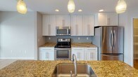 This single-floor residence offers a thoughtfully designed layout with a clean, contemporary aesthetic throughout. The kitchen serves as a highlight of the space, showcasing white shaker-style cabinetry extending to the ceiling, warm gold-toned granite countertops, and a full suite of stainless steel appliances including a range, over-the-range microwave, and refrigerator. A central island with an undermount sink and pendant lighting above provides additional workspace and a natural gathering point. Recessed lighting and light wood-style flooring complete the kitchen's polished look.

The bedroom areas feature neutral-toned carpeting and bright white walls, creating a calm and versatile backdrop. Large windows in the main living area allow natural light to fill the room, offering views to the outdoors. One bedroom includes a dedicated walk-in closet equipped with a hanging rod, providing organized storage space.

The bathroom is efficiently arranged, featuring a frameless glass-enclosed shower with brushed nickel fixtures, a toilet, and direct access to the walk-in closet. A wall-mounted flush light fixture provides even illumination. Throughout the unit, HVAC vents and clean white baseboards contribute to a cohesive, well-maintained finish.