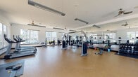 Located at 2200 Bellamy Dr in Greenville, NC, this fitness center offers a well-appointed, single-floor facility designed to support a wide range of workout routines. The space is characterized by its bright, open layout, featuring large windows along multiple walls that flood the interior with abundant natural light and provide views of the surrounding community. The durable rubberized flooring throughout provides both safety and comfort during exercise.

The strength training area is generously equipped with a variety of cable machines, multi-function weight stations, and a full dumbbell rack, complemented by floor space for free-weight and bodyweight exercises. Full-length mirrors line one wall, enhancing the sense of space and allowing users to monitor their form. Ceiling fans and industrial-style pendant lighting ensure comfort and visibility throughout.

The cardio section features multiple treadmills, elliptical machines, and stationary bikes, positioned near large windows to provide an engaging outdoor view during workouts. A dedicated stretching and flexibility area includes yoga mats and foam rollers stored on a rack, offering a functional space for warm-ups and cool-downs. The facility's clean, white walls and open floor plan create an energizing and motivating environment suitable for users of all fitness levels.
