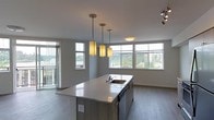 This single-floor space showcases a thoughtfully designed open-concept layout that seamlessly connects the kitchen and living areas. At the heart of the space sits a sleek kitchen island with a light quartz countertop and an integrated sink, offering both functionality and a natural gathering point. Warm pendant lights hang in a staggered arrangement above the island, providing focused task lighting while adding a stylish visual element to the space.

The kitchen cabinetry features a contemporary grey-toned finish, complementing the neutral color palette found throughout. Stainless steel appliances, including a built-in dishwasher, contribute to the modern aesthetic.

Large horizontal windows line the perimeter of the open living area, flooding the space with abundant natural light and framing pleasant views of surrounding greenery and the urban skyline beyond. A sliding door leads to an outdoor balcony, further enhancing the connection to the outside environment.

Light-toned hardwood-style flooring runs throughout, adding warmth and continuity across the open layout. The clean white ceilings and soft grey walls create a calm, neutral backdrop that makes the space feel bright, airy, and versatile.
