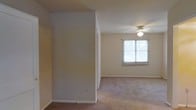 This single-floor space offers a straightforward and functional layout with a clean, neutral aesthetic throughout. The interior features wall-to-wall carpeting in a warm beige tone, providing a comfortable underfoot feel that carries through the rooms. The walls are finished in a soft, neutral color palette, creating a calm and versatile backdrop well-suited to a variety of furnishing styles.

A central room divider or partial wall adds a subtle sense of structure to the open layout, defining areas within the space without fully enclosing them. The ceiling is fitted with a ceiling fan and integrated light fixture, offering both practical lighting and air circulation. A window positioned along one wall allows natural light to filter into the room, brightening the interior and giving a sense of openness despite the modest proportions.

The overall ambiance is understated and move-in ready, with clean lines and a simple design that allows occupants to personalize the space to their needs. The neutral finishes and carpeted floors give the space a cozy, livable quality, while the straightforward layout ensures ease of use and adaptability for everyday living.