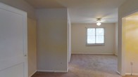 This single-floor space offers a straightforward and functional layout with a clean, neutral aesthetic throughout. The interior features wall-to-wall carpeting in a warm beige tone, providing a comfortable underfoot feel that carries through the rooms. The walls are finished in a soft, neutral color palette, creating a calm and versatile backdrop well-suited to a variety of furnishing styles.

A central room divider or partial wall adds a subtle sense of structure to the open layout, defining areas within the space without fully enclosing them. The ceiling is fitted with a ceiling fan and integrated light fixture, offering both practical lighting and air circulation. A window positioned along one wall allows natural light to filter into the room, brightening the interior and giving a sense of openness despite the modest proportions.

The overall ambiance is understated and move-in ready, with clean lines and a simple design that allows occupants to personalize the space to their needs. The neutral finishes and carpeted floors give the space a cozy, livable quality, while the straightforward layout ensures ease of use and adaptability for everyday living.