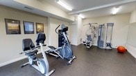 The fitness center is a single-level space designed to provide residents with convenient access to essential exercise equipment. The room features durable, speckled rubber flooring that offers both safety and easy maintenance, ideal for a high-use workout environment. Bright fluorescent overhead lighting ensures the space is well-lit throughout, creating a clear and energizing atmosphere conducive to exercise.

The layout accommodates a variety of workout options. Cardio equipment includes a recumbent bike and an elliptical trainer, both positioned to allow comfortable use without overcrowding. A multi-station cable and weight machine occupies the far end of the room, offering users options for comprehensive strength training. A freestanding dumbbell rack provides additional versatility for free weight exercises.

The walls are painted in a neutral cream tone, keeping the space feeling open and clean. Motivational fitness posters are displayed along one wall, adding character to the room. A closet door near the back suggests additional storage within the space. The overall design prioritizes functionality, offering a straightforward and accessible environment for daily fitness routines.