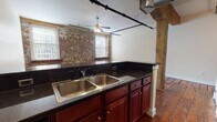This single-floor space showcases a compelling mix of historic industrial character and functional modern updates. Throughout the unit, rich reclaimed hardwood plank flooring sets a warm, rustic tone, complemented by exposed original wood beam columns that serve as striking architectural focal points. Black iron piping runs visibly along the ceilings, reinforcing the industrial aesthetic alongside ceiling fans that promote airflow throughout the open layout.

The kitchen features rich cherry wood cabinetry, dark granite countertops, a double stainless steel sink, and stainless steel appliances including a refrigerator, electric range, and microwave. An exposed brick wall with two windows brings natural light and textural contrast to the cooking area. The kitchen opens directly to a generous living area, creating a cohesive, open-concept flow ideal for everyday living.

The bedrooms continue the hardwood flooring and neutral wall tones, offering clean, versatile spaces. One room includes a dedicated walk-in closet with wire shelving and the same warm hardwood underfoot. Two bathrooms serve the unit: one full bath with a tub/shower combo, tile flooring, and a cherry vanity, and a second half bath with matching finishes. The overall ambiance is warm, characterful, and rooted in the building's architectural history.