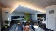 This single-floor space showcases a thoughtfully designed modern lounge area that balances open-plan living with subtle spatial definition. The interior features a striking suspended ceiling element with integrated indirect lighting, which casts a warm, ambient glow against the exposed concrete ceiling above, creating a layered architectural effect that adds depth and visual interest to the space.

A distinctive vertical green slat partition serves as a bold design focal point, dividing the space while maintaining visual connectivity between zones. This vibrant divider introduces a natural color element that contrasts effectively with the otherwise neutral palette of cool greys and whites throughout the space.

The lounge area is furnished with sleek, angular brown leather chairs accented with olive-toned cushions, arranged around cylindrical side tables in varying heights and metallic finishes. The multi-tonal grey carpeting adds texture underfoot and complements the overall contemporary aesthetic.

In the background, a secondary area features long communal tables with matching seating, suggesting a collaborative or dining function. The overall ambiance is polished, modern, and professional, making the space well-suited for both relaxation and social interaction.
