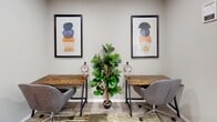 Located at 2096 E Main St in Spartanburg, South Carolina, this single-floor space offers a thoughtfully curated environment that balances functionality with contemporary style. The interior features neutral gray walls that serve as a versatile backdrop for bold, colorful accents throughout.

A welcoming lounge area anchors one section of the space, highlighted by a vibrant yellow tufted sofa positioned beneath a trio of striking circular abstract artworks rendered in yellow and purple tones. Complementing this area is a plush gray tufted settee positioned near a large window dressed with white faux-wood blinds, allowing ample natural light to filter in while offering views of the outdoors. A tall fiddle-leaf fig plant adds a lush, organic element to this corner.

A dedicated work area features two rustic wood-topped desks paired with modern gray upholstered task chairs, each equipped with headphones and framed by contemporary geometric artwork. A large indoor plant between the desks enhances the refreshing, focused atmosphere.

Additional seating is provided by a pair of low-profile, cushioned lounge chairs positioned near a set of bi-fold closet doors, offering a relaxed retreat within the space. The flooring throughout features a patterned area rug layered over hard flooring, tying the various zones together cohesively. Overall, the space exudes a modern, creative, and professionally appointed character.