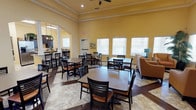 This single-story communal space is designed to foster social interaction and comfort. The main dining area is characterized by warm yellow walls, elegant crown molding, and elevated ceilings that lend an open, airy feel. A ceiling fan overhead provides both function and a finishing architectural touch. The flooring features a distinctive combination of dark hardwood and neutral tile laid in a geometric pattern, adding visual interest throughout the space.

Multiple square dining tables, each surrounded by dark wood chairs with tan cushioned seats, are arranged throughout the room, offering flexible seating for individuals or groups. Natural light filters in through several white-shuttered windows along one wall, brightening the cheerful interior.

A decorative arched doorway framed by white columns connects the main dining area to an adjacent kitchen or café-style space, visible in the background with dark cabinetry and pendant lighting. A small lounge area with upholstered seating sits near the windows, providing a relaxed alternative to the dining tables. Together, these elements create a welcoming, multi-functional environment suited for dining, socializing, and everyday community activities.