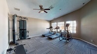 Located at 3245 E University Ave in Las Cruces, New Mexico, this fitness center offers a well-equipped, single-floor workout space designed to support a range of exercise needs. The room features durable, patterned carpet tile flooring throughout, providing comfort underfoot during workouts. Neutral-toned walls and a textured ceiling contribute to a clean, functional aesthetic.

The cardio area is outfitted with multiple Matrix treadmills and an elliptical trainer, all positioned along a bank of windows that allow natural light to fill the space and provide views of the outdoor surroundings. A ceiling fan overhead aids in air circulation, keeping the environment comfortable during use.

A dedicated strength training corner houses a Matrix functional trainer, a versatile cable machine that supports a wide variety of resistance exercises. The equipment is thoughtfully arranged to allow users ample space to move freely between stations.

Access to the fitness center is through elegant French doors with glass panes, connecting the gym to an adjacent space. An additional standard door provides further entry and exit options. The overall layout is open and easy to navigate, making it a practical and inviting environment for residents seeking convenient access to fitness facilities in Las Cruces.