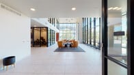 This single-floor commercial lobby is designed with a clean, contemporary aesthetic that immediately conveys a professional and welcoming atmosphere. The centerpiece of the space is a dramatic wall of floor-to-ceiling windows framed in dark steel, flooding the interior with abundant natural light and offering clear views of the surrounding outdoor landscape and neighboring structures. Large-format light gray porcelain tile flooring extends throughout, contributing to a bright and polished finish underfoot.

At the heart of the lobby, a curated lounge area features curved amber-hued upholstered chairs arranged around a slender round table, anchored by a low-pile area rug — offering a comfortable and stylish gathering spot. Adjacent to the lounge, a glass-walled office or meeting room enclosed in black steel framing provides a semi-transparent private workspace visible from the main floor.

Along one wall, dual large-format digital display screens present property and amenity content, complemented by sleek modern planters positioned nearby. Automatic glass entry doors with dark metal frames provide accessible access to the building. The overall ambiance is polished and professional, balancing openness and transparency with purposeful zoning — making it well-suited for a high-end office, residential leasing center, or corporate headquarters entrance.