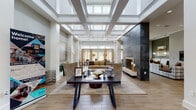 This elegantly designed single-floor communal space makes an immediate impression with its grand, coffered ceiling fitted with expansive skylights that flood the interior with natural light. Structural columns frame the open central hall, reinforcing the sense of volume and architectural refinement. Light wood-look flooring runs throughout, providing a warm, cohesive foundation across all areas.

The main lounge area serves as the focal point, anchored by a floor-to-ceiling dark stone fireplace that adds dramatic contrast to the otherwise neutral palette. Comfortable seating arrangements, including upholstered chairs and sofas, are thoughtfully positioned around a central table, creating an inviting gathering environment. Curated artwork and greenery enhance the refined ambiance throughout.

A secondary lounge area features built-in banquette seating, pendant globe lighting, and a wall-mounted television, offering a more relaxed, intimate setting. Distinct accent walls in muted sage green add depth and visual interest to this zone.

A dedicated beverage station, highlighted by a striking multicolored mosaic tile backsplash, includes a coffee machine, beverage refrigerator, and granite countertops with white cabinetry. Nearby, glass-top bistro tables positioned beside floor-to-ceiling draped doors open directly to an outdoor pool area, seamlessly connecting interior comfort with outdoor amenity access.