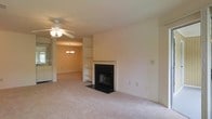 This single-floor space offers a functional and comfortable layout with a seamless flow between the living and dining areas. The living room serves as the focal point, centered around a classic fireplace with a light-toned surround and dark hearth, adding warmth and character to the room. Built-in shelving flanks the fireplace, providing practical storage and display space. The room is finished with plush wall-to-wall carpeting in a neutral tone, contributing to a soft, inviting atmosphere throughout.

A ceiling fan overhead offers both practical ventilation and casual lighting for the living area. The open layout connects to the dining area, which is illuminated by an elegant chandelier, creating a natural transition between spaces. A glimpse into what appears to be a kitchen is visible beyond the dining area, suggesting a well-connected floor plan ideal for everyday living and entertaining.

The walls throughout are painted in a clean, neutral tone, offering a versatile backdrop that complements a variety of interior styles. Overall, this space combines classic details with a straightforward, open design.