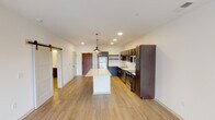 This single-floor unit is designed with a clean, contemporary aesthetic throughout. The kitchen serves as a central highlight, showcasing rich dark cabinetry paired with light quartz countertops, a white subway tile backsplash, and a full suite of stainless steel appliances including a refrigerator, electric range, microwave, and dishwasher. A generously sized central island with a bright quartz surface anchors the space, illuminated by a pair of modern pendant lights that add a stylish focal point. Open shelving and a classic barn door add character and functional storage to the kitchen area.

The living area is bright and open, featuring light hardwood-style flooring that flows seamlessly throughout the main spaces. Natural light enters through large windows and a glass-panel door that provides direct access to an outdoor balcony, enhancing the sense of openness. The bedroom is carpeted for comfort and receives natural light through a large window with exterior views.

The bathroom is finished with white subway tile in the shower surround, complemented by light hardwood-style flooring and clean white walls. Shaker-style interior doors and recessed lighting throughout contribute to the unit's cohesive, modern design.