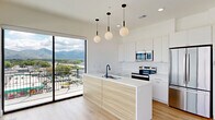 This contemporary 2-bedroom, 1-bathroom residence offers a thoughtfully designed single-floor layout bathed in natural light. The open-concept kitchen serves as the heart of the home, featuring a wood-paneled island with a white quartz countertop and undermount sink, sleek white cabinetry extending to the ceiling, stainless steel appliances, and a trio of globe pendant lights. A large sliding glass door connects the kitchen directly to a spacious private balcony, where sweeping panoramic views of the surrounding mountains and cityscape take center stage beneath an open sky. Light-toned hardwood flooring flows seamlessly throughout the entire residence, enhancing the bright, airy atmosphere. The primary bedroom is generously sized, equipped with a ceiling fan and floor-to-ceiling windows that frame expansive outdoor views. The second bedroom also features a ceiling fan and access to the balcony through a sleek glass door. A dedicated closet with built-in shelving provides practical storage. The bathroom is finished with a warm wood vanity, white quartz countertop, a large wall mirror, and a marble-tiled walk-in shower. Clean white walls and recessed lighting throughout contribute to a crisp, modern aesthetic.