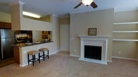 This single-floor residence offers a comfortable and functional layout highlighted by several notable design features. The living area serves as the focal point of the space, centered around a classically styled white-mantled fireplace that adds warmth and character to the room. Soft, neutral-toned carpeting runs throughout, complementing the warm beige walls accented by white crown molding and baseboards. A ceiling fan with a light fixture is positioned above, providing both air circulation and ambient lighting.

Adjacent to the living area, a pass-through kitchen window opens to a breakfast bar equipped with two cushioned bar stools, creating a convenient casual dining area. The kitchen features dark granite-style countertops and wood-toned cabinetry, visible through the pass-through opening. Overhead lighting illuminates the kitchen workspace effectively.

A closet door is visible near the center of the space, offering practical storage. The overall ambiance of this residence is understated and inviting, with a cohesive neutral color palette that creates a clean, livable environment throughout.