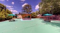 Located at 8805 Summit Place Dr in Sandy Springs, Georgia, this outdoor recreational space offers a generous rubber-surfaced court area finished in a distinctive green coating, providing ample room for a variety of activities. The grounds are thoughtfully appointed with multiple picnic tables shaded by teal umbrellas, offering comfortable gathering spots throughout the space. A wooden pergola structure anchors one end of the court, adding architectural character and a defined social area. Tiered stone and wood retaining walls create natural amphitheater-like seating along the perimeter, blending seamlessly with the surrounding landscape. Tall wooden plank fencing and rail fencing define the boundaries, providing both privacy and a warm, natural aesthetic. Pole-mounted lighting ensures the space remains functional into the evening hours. Lush, mature trees and dense greenery frame the entire area, creating a secluded, park-like atmosphere. Benches are positioned throughout for additional seating. The landscaping includes raised planting beds with evergreen shrubs and trees, enhancing the verdant setting. Open grassy areas beyond the court add depth to the outdoor environment, contributing to the overall sense of open, natural space in this well-equipped community recreational area.