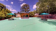 Located at 8805 Summit Place Dr in Sandy Springs, Georgia, this outdoor recreational space offers a generous rubber-surfaced court area finished in a distinctive green coating, providing ample room for a variety of activities. The grounds are thoughtfully appointed with multiple picnic tables shaded by teal umbrellas, offering comfortable gathering spots throughout the space. A wooden pergola structure anchors one end of the court, adding architectural character and a defined social area. Tiered stone and wood retaining walls create natural amphitheater-like seating along the perimeter, blending seamlessly with the surrounding landscape. Tall wooden plank fencing and rail fencing define the boundaries, providing both privacy and a warm, natural aesthetic. Pole-mounted lighting ensures the space remains functional into the evening hours. Lush, mature trees and dense greenery frame the entire area, creating a secluded, park-like atmosphere. Benches are positioned throughout for additional seating. The landscaping includes raised planting beds with evergreen shrubs and trees, enhancing the verdant setting. Open grassy areas beyond the court add depth to the outdoor environment, contributing to the overall sense of open, natural space in this well-equipped community recreational area.